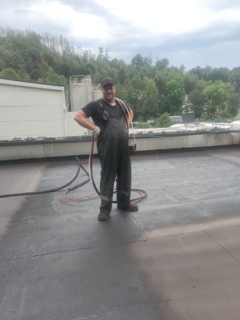 Een persoon in overall en pet staat op een dak met een slang in zijn hand, vakkundig bezig met waterafdichting. De achtergrond toont bomen, een gebouw en een bewolkte lucht.