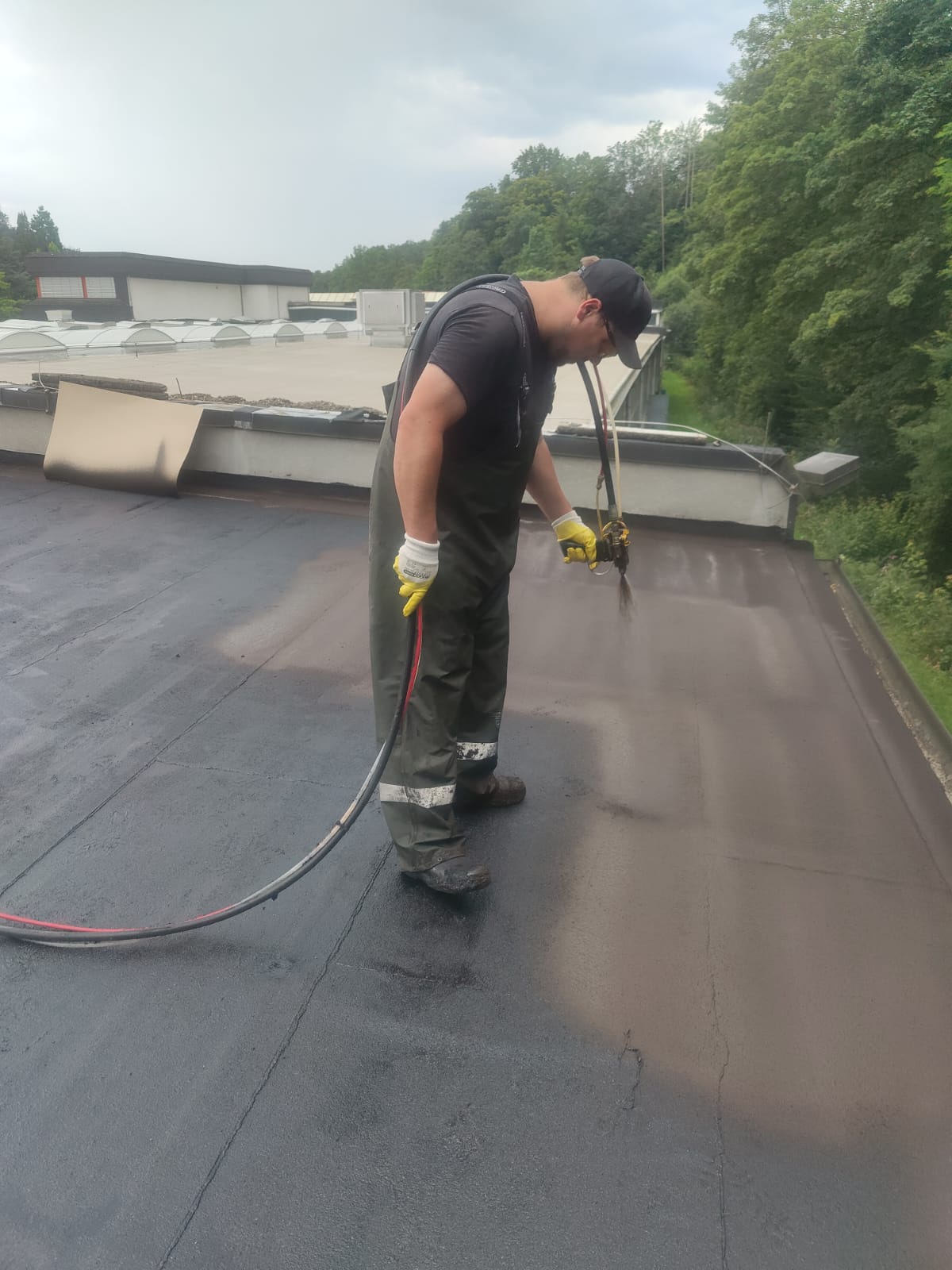 Een persoon in werkkleding en handschoenen brengt vakkundig een dakafdichtinglaag aan op een plat dak met behulp van een speciaal gereedschap. De omgeving is weelderig met bomen onder een bewolkte lucht, wat een ideale setting creëert voor nauwkeurig waterafdichtingwerk.