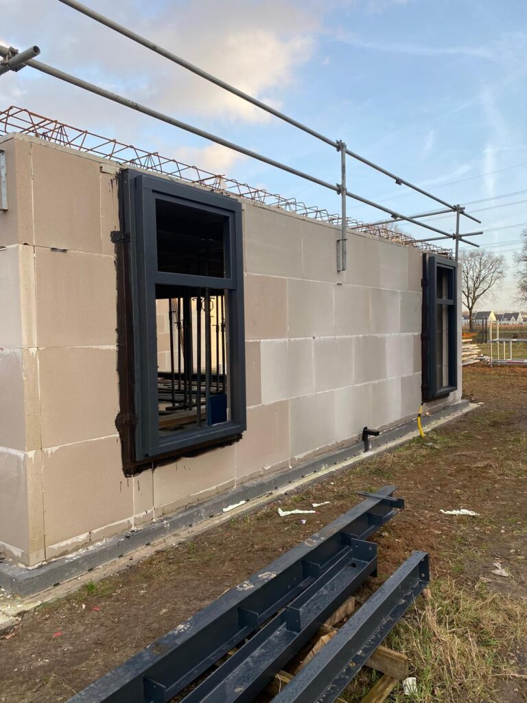 Een gedeeltelijk gebouwd gebouw heeft een betonnen blokmuur met twee grote raamopeningen in een zwart frame. Steigers steken erboven uit, wat zorgt voor luchtdicht bouwen. Metalen balken liggen op het gras op de voorgrond onder een helderblauwe lucht.