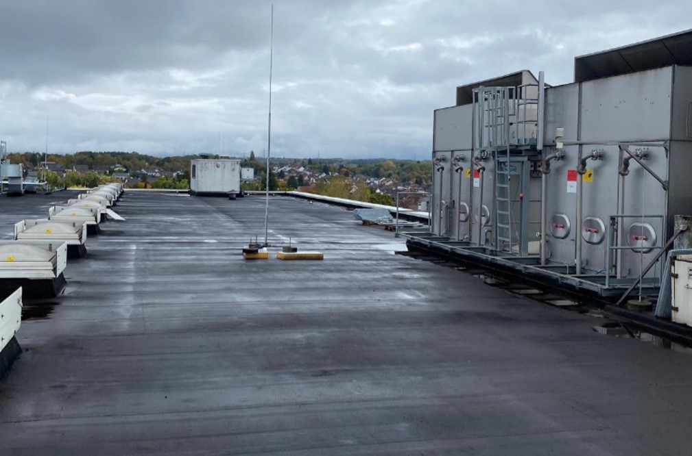 Een dakaanzicht toont industriële HVAC-units en -apparatuur onder een bewolkte lucht. Het platte dak, beschermd door dakafdichting, is bedekt met een donker materiaal, met gebouwen en bomen op de verre achtergrond.