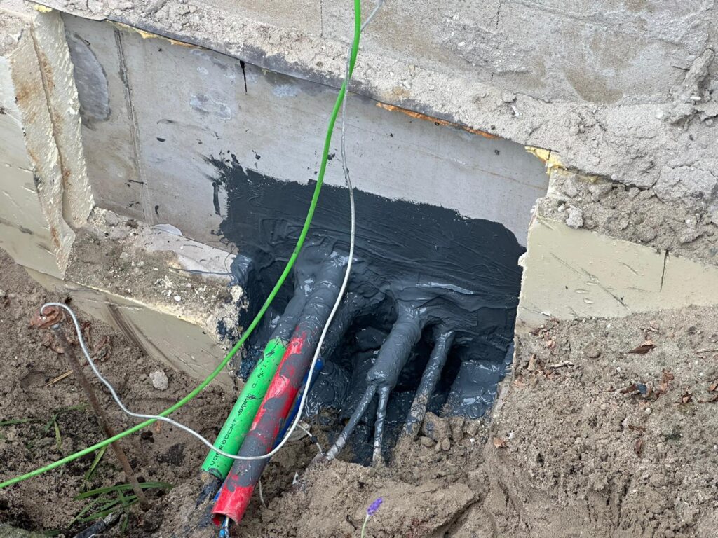 Kabels omhuld met een zwarte betonafdichting komen uit een betonnen muur in een bouwgebied. Twee groene en rode beschermhoezen zijn naast de kabels geplaatst, omgeven door vuil en gedeeltelijk puin.